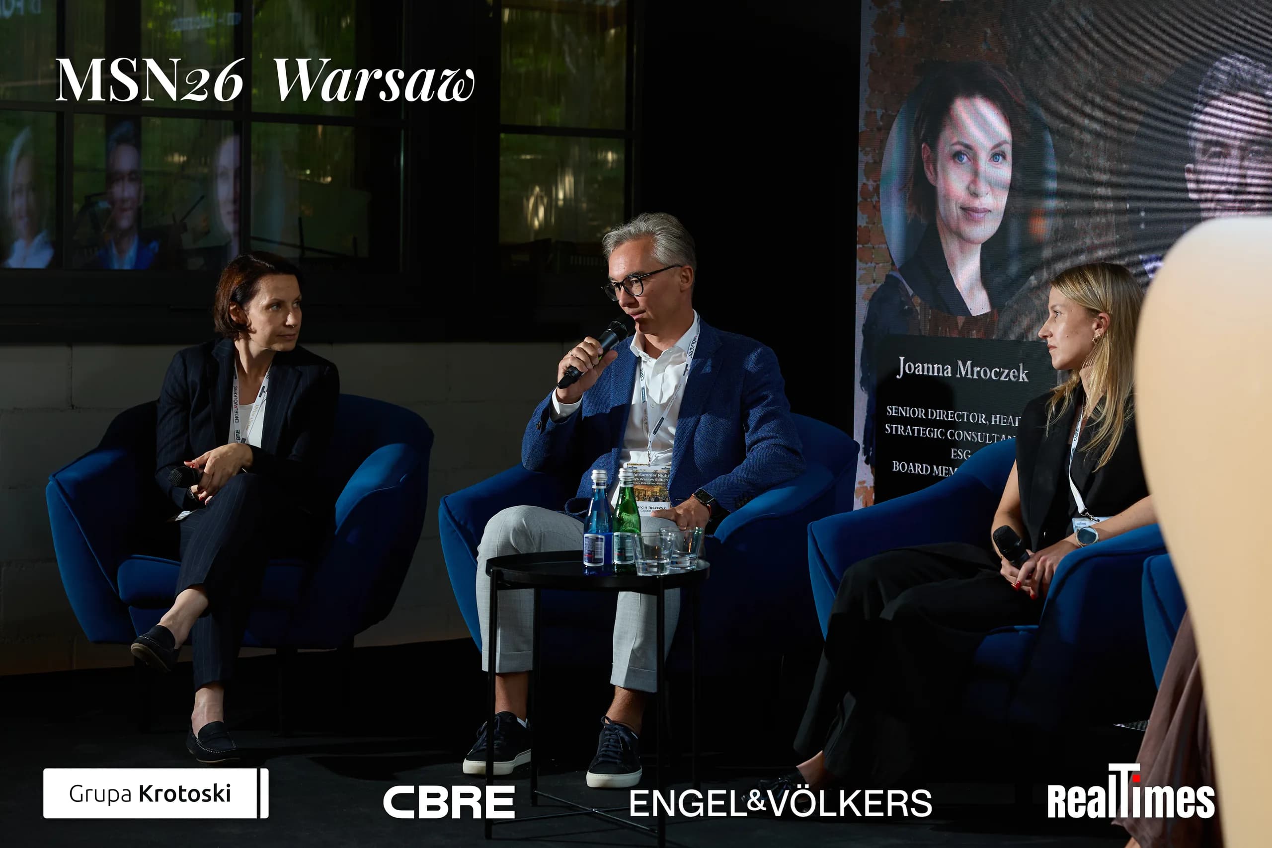 Mid-Summer Night 2026 at B'Fort, Warsaw: man with mic + panelists, Mroczek card behind