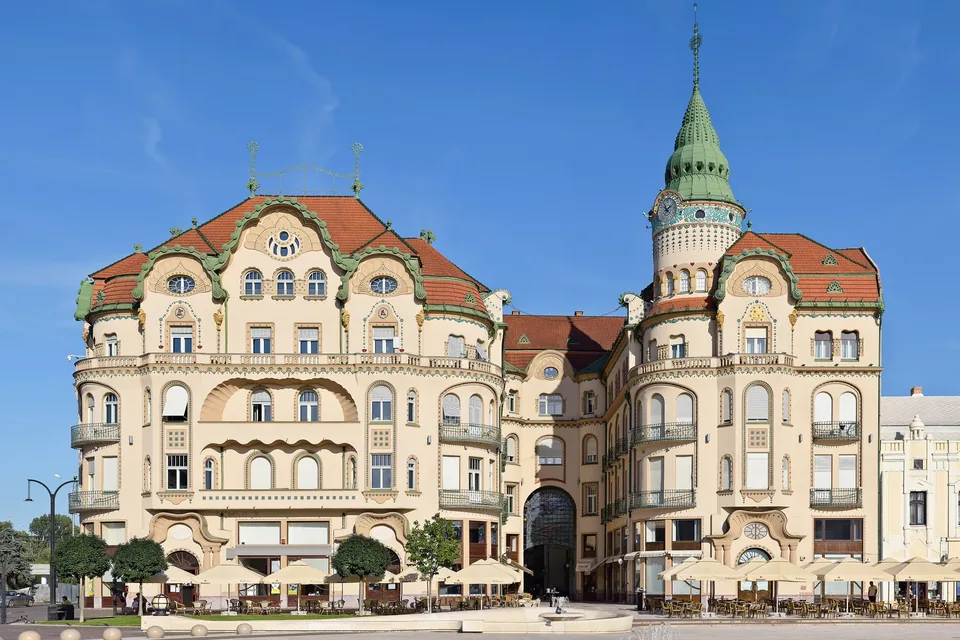 The Black Eagle Palace on Piața Unirii in Oradea, a cream Secession-era building with green ornament, red tiled roofs and a green spired clock tower, café umbrellas along the square under a clear blue sky.