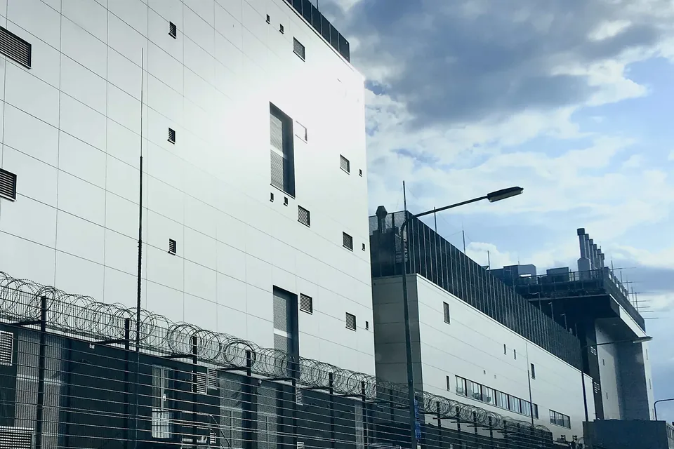 A windowless data-centre building in Frankfurt's Ostend, razor-wire security fencing along its base and cooling plant with exhaust stacks on the roof of the hall behind it.