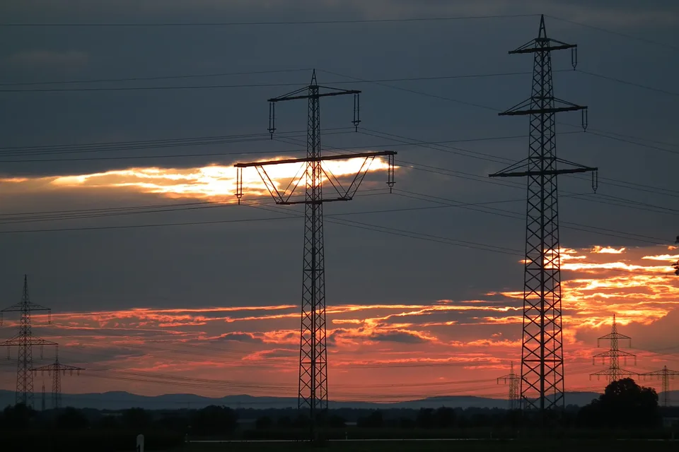 High-voltage transmission pylons in silhouette against a red and orange sunset over flat farmland in northern Baden-Württemberg, more pylons receding towards distant hills.