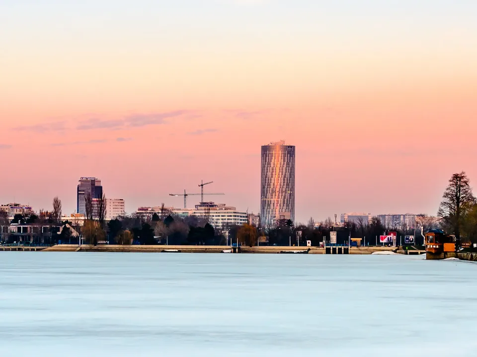 Bucharest's Sky Tower catching the last warm light at sunset, rising above the Floreasca office district and two tower cranes, seen across the frozen surface of Herăstrău lake under a pink and amber sky.