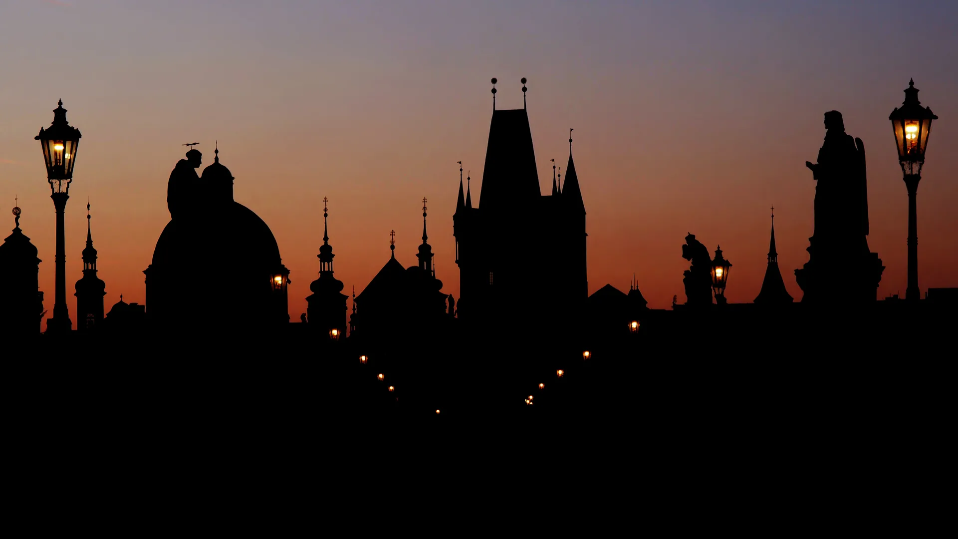 Silhouettes of Prague's towers, domes and Charles Bridge statues against an orange dawn sky, street lamps still lit.