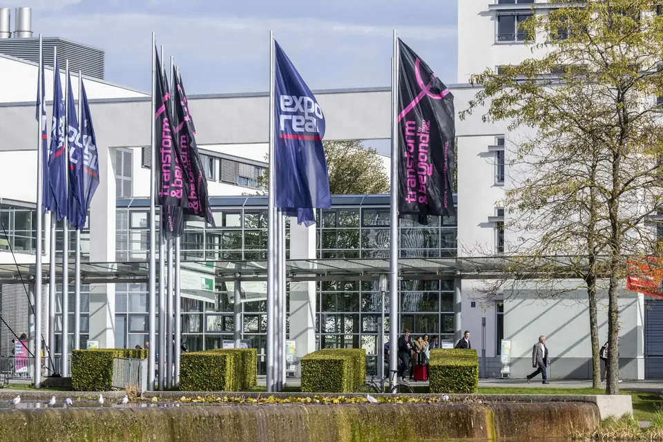 Flags outside the west entrance of Messe München during EXPO REAL 2025, with the glass entrance hall and a few visitors walking past.