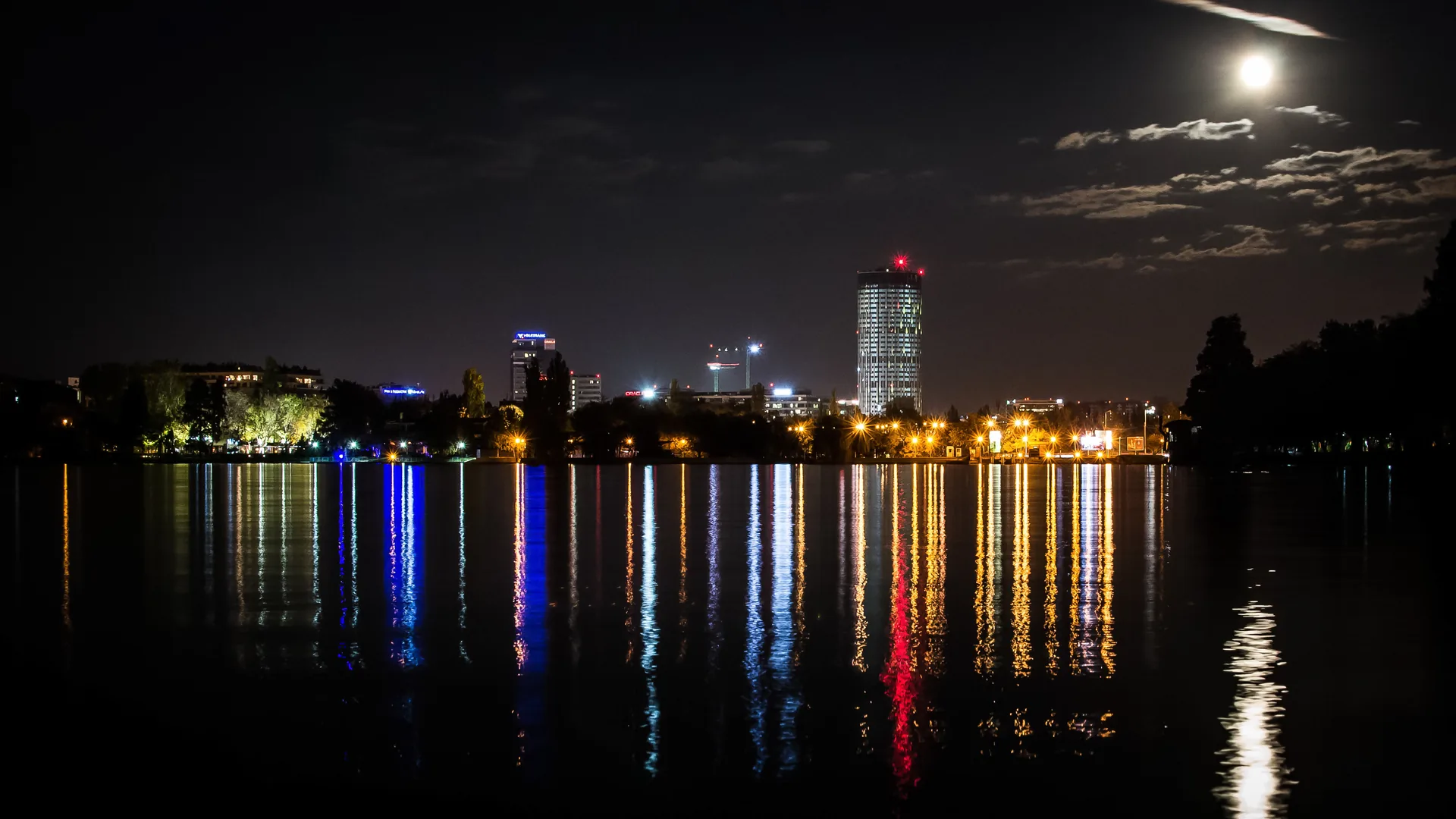 Bucharest's Sky Tower and the northern office district lit at night across Herăstrău lake, the lights reflected in the water under a full moon.