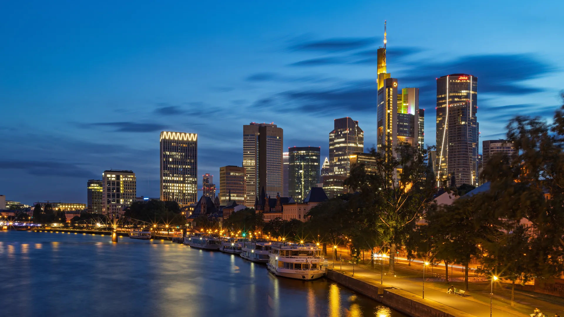 Frankfurt's lit banking towers above the River Main at blue hour, river boats moored along the embankment in the foreground.