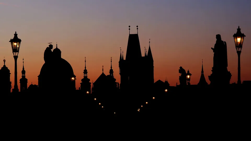 Silhouettes of Prague's towers, domes and Charles Bridge statues against an orange dawn sky, street lamps still lit.