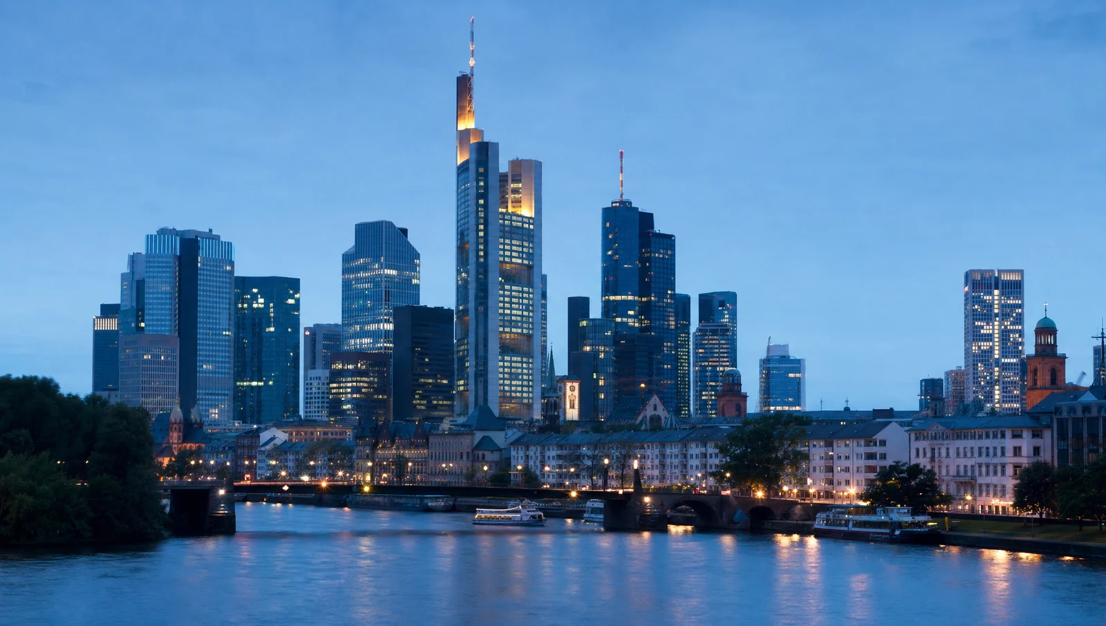 Frankfurt banking-district skyline at blue hour seen across the river Main, the lit crown and antenna of the Commerzbank Tower at the centre of the cluster, the Alte Bruecke and river boats below