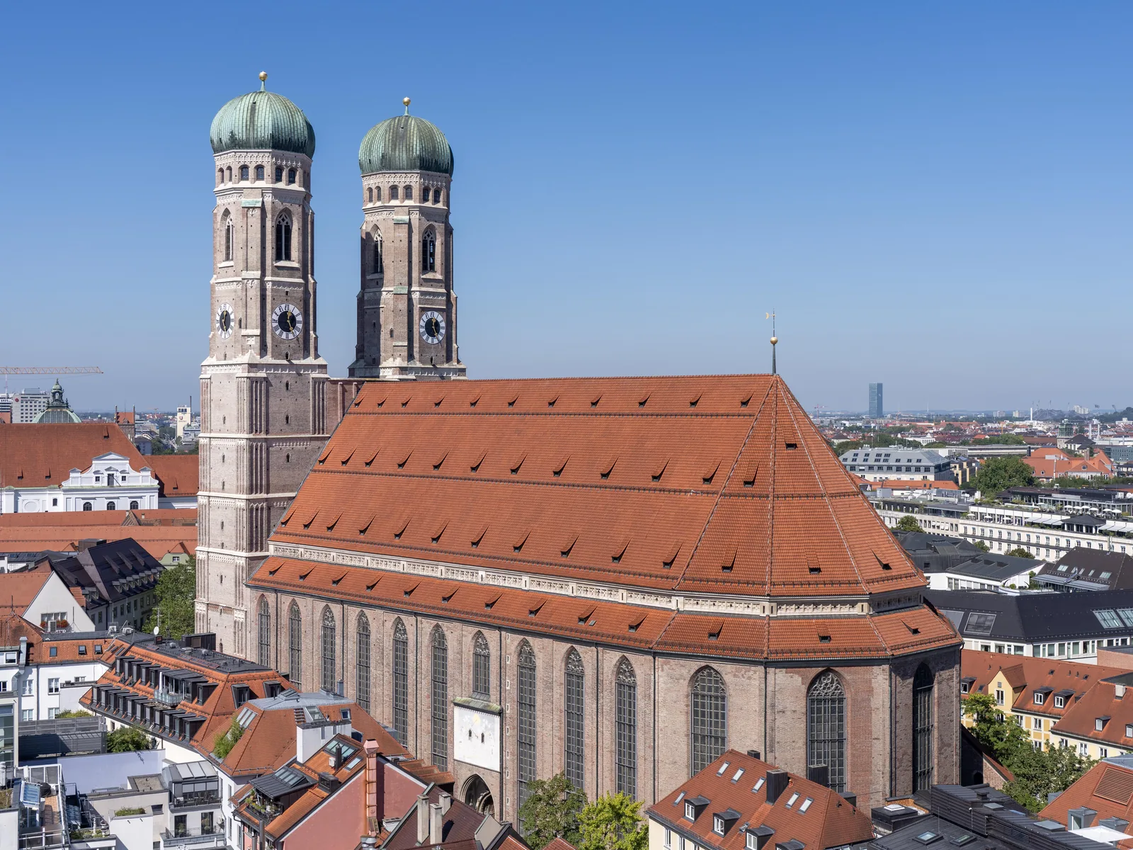 The twin domed towers and steep red roof of Munich's Frauenkirche above the old-town rooftops on a clear day, with a distant office tower rising on the horizon.